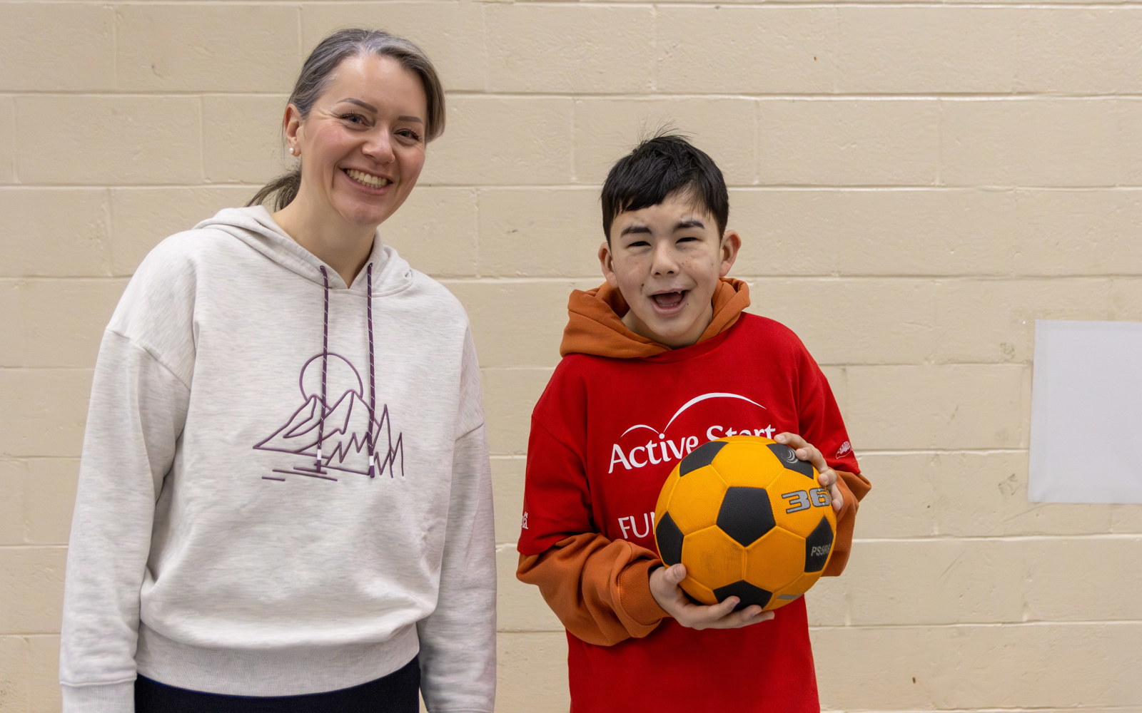 A young athlete with intellectual disabilities stands next to his coach while holding a soccer ball.