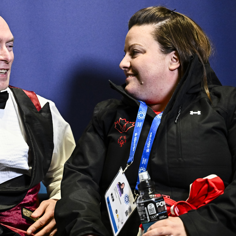 A figure skater holds a small bouquet of flowers and teddy bear while sitting and smiling with his coach.
