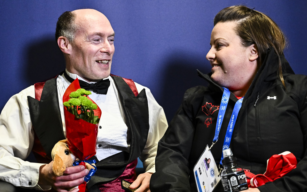A figure skater holds a small bouquet of flowers and teddy bear while sitting and smiling with his coach.