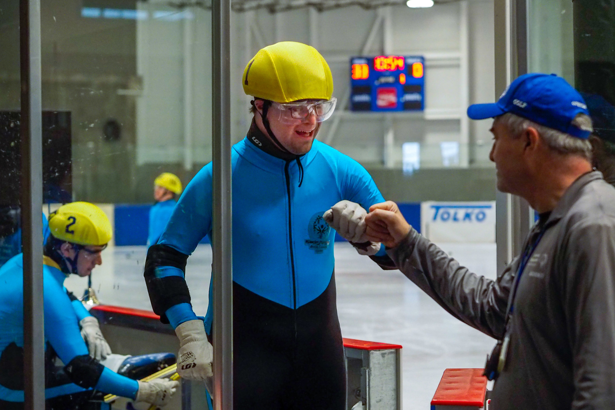 A speed skating athlete in a yellow helmet fist bumps his coach as he exits the ice.