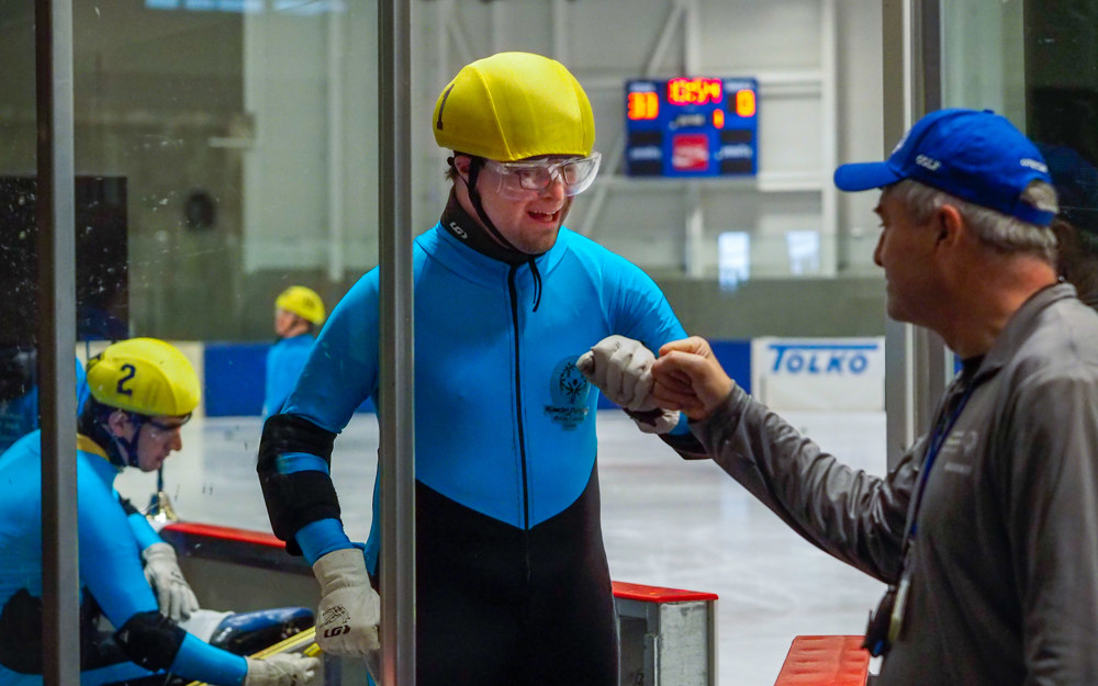 A speed skating athlete in a yellow helmet fist bumps his coach as he exits the ice.