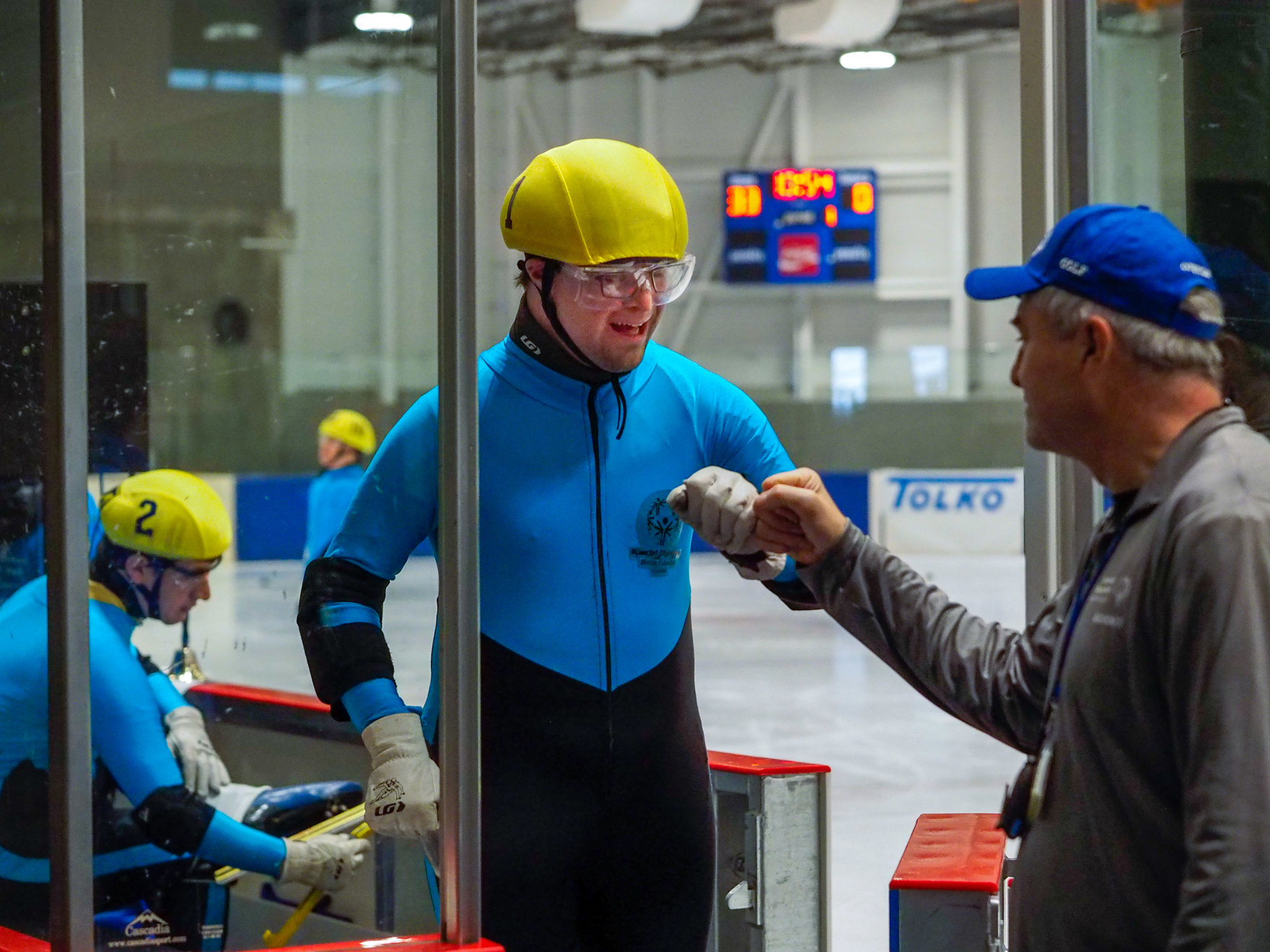 A speed skating athlete in a yellow helmet fist bumps his coach as he exits the ice.