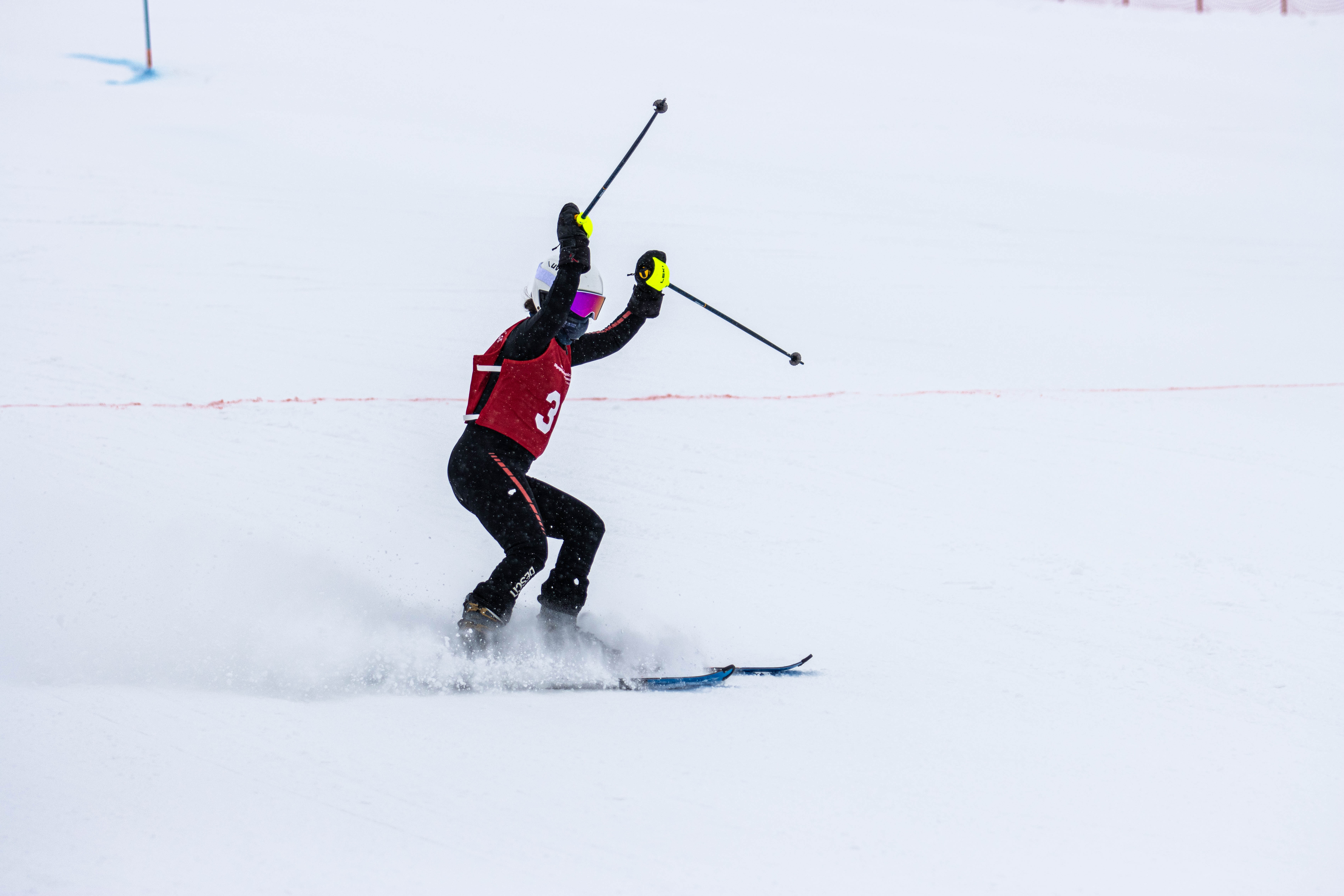 Special Olympics BC alpine skiing athlete celebrates at the finish line with their hands and poles raised in the air and a cloud of snow off their skis as they come to a stop.