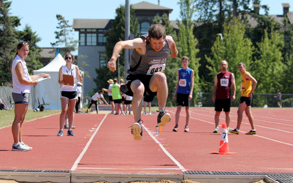Special Olympics BC athlete competing in long jump is shown mid-jump before landing in the sand.