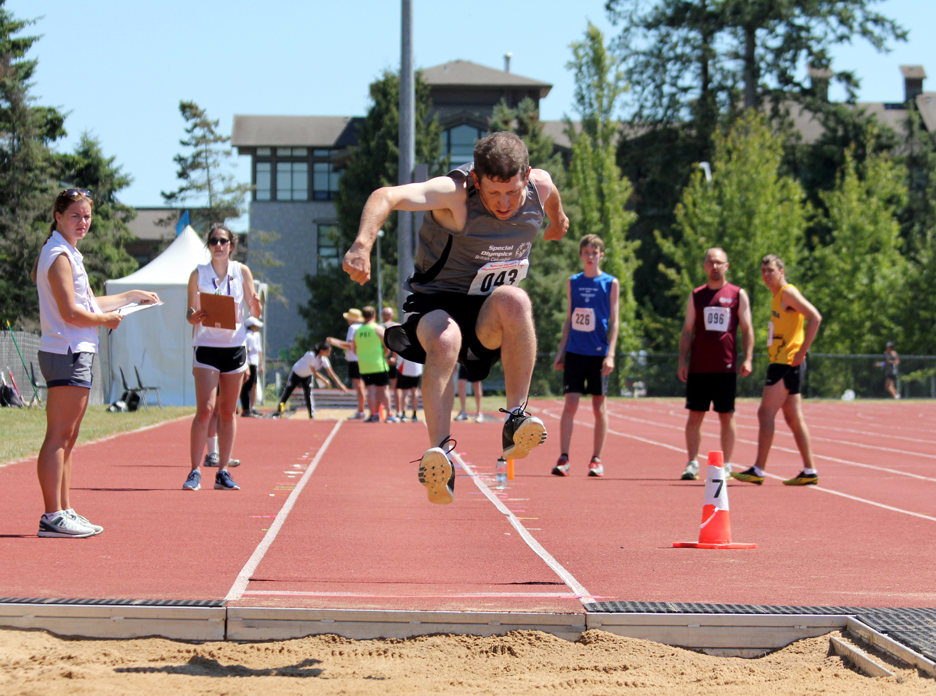 Special Olympics BC athlete competing in long jump is shown mid-jump before landing in the sand.