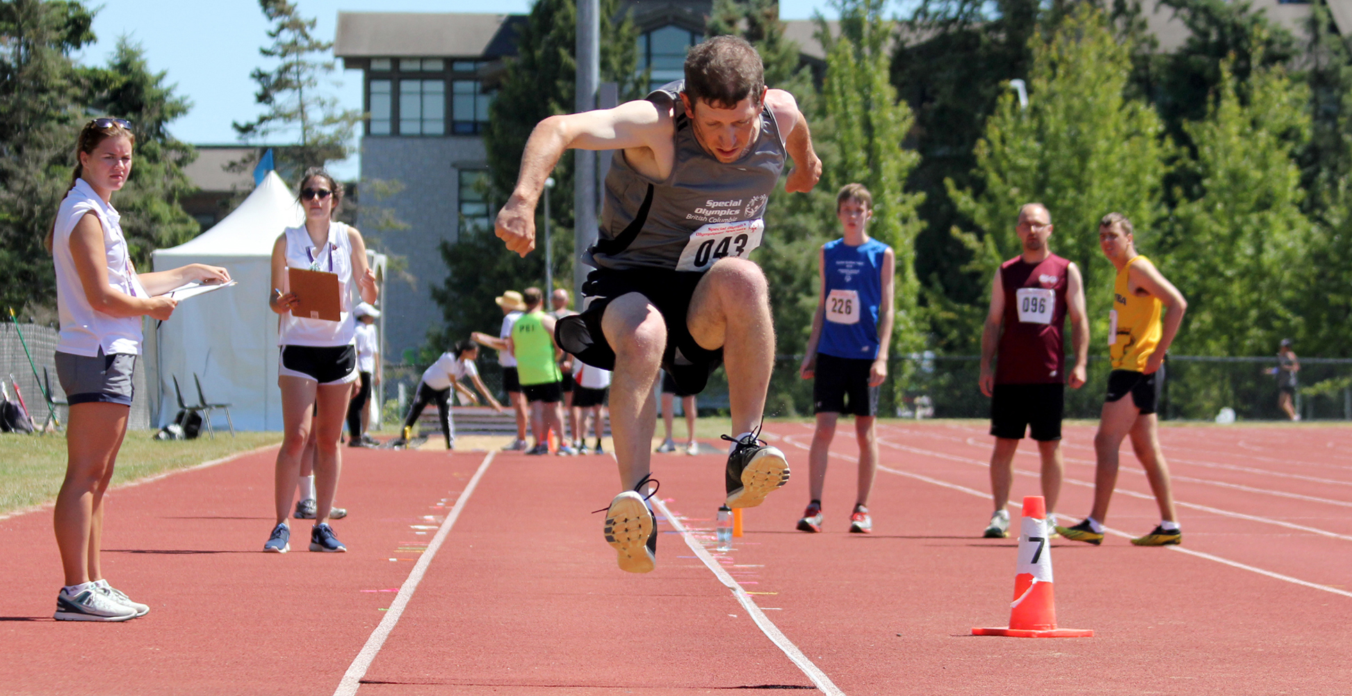 Special Olympics BC athlete competing in long jump is shown mid-jump before landing in the sand.