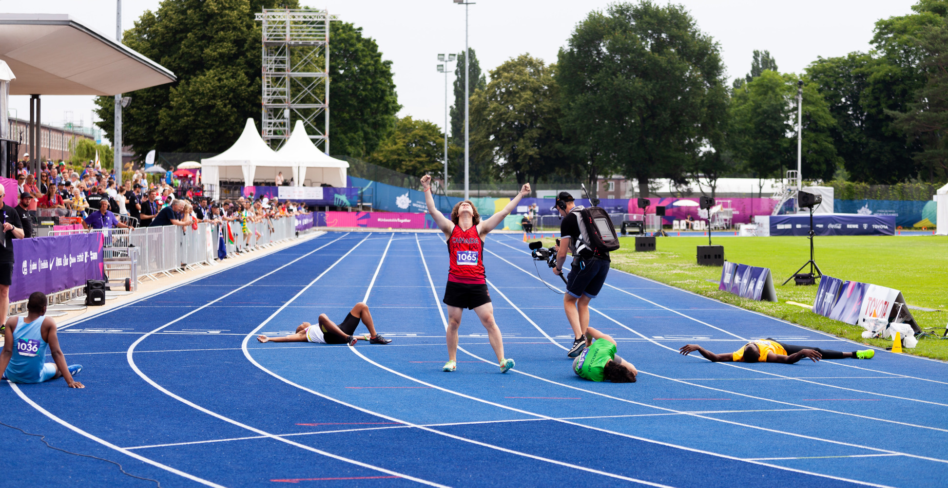 A triumphant Special Olympics BC athlete at the end of their race with their arms raised as fellow competitors lie exhausted on the track around him.