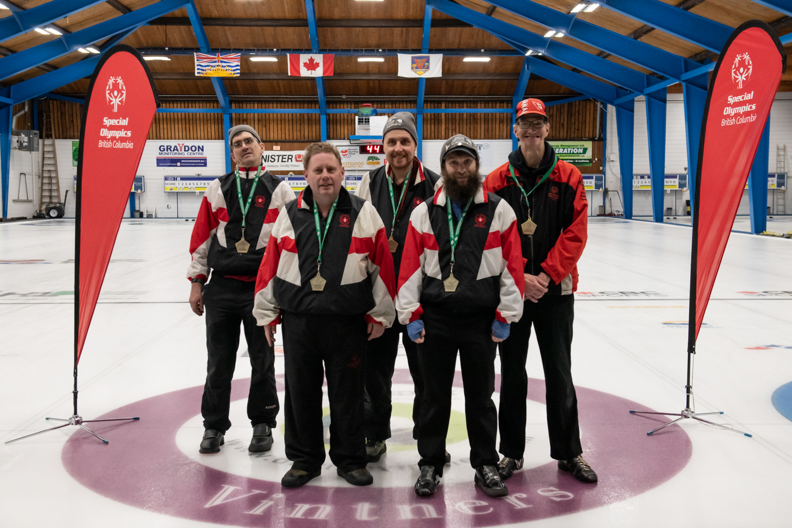 A proud team of Special Olympics BC curling athletes poses together with their coach with medals around their necks.
