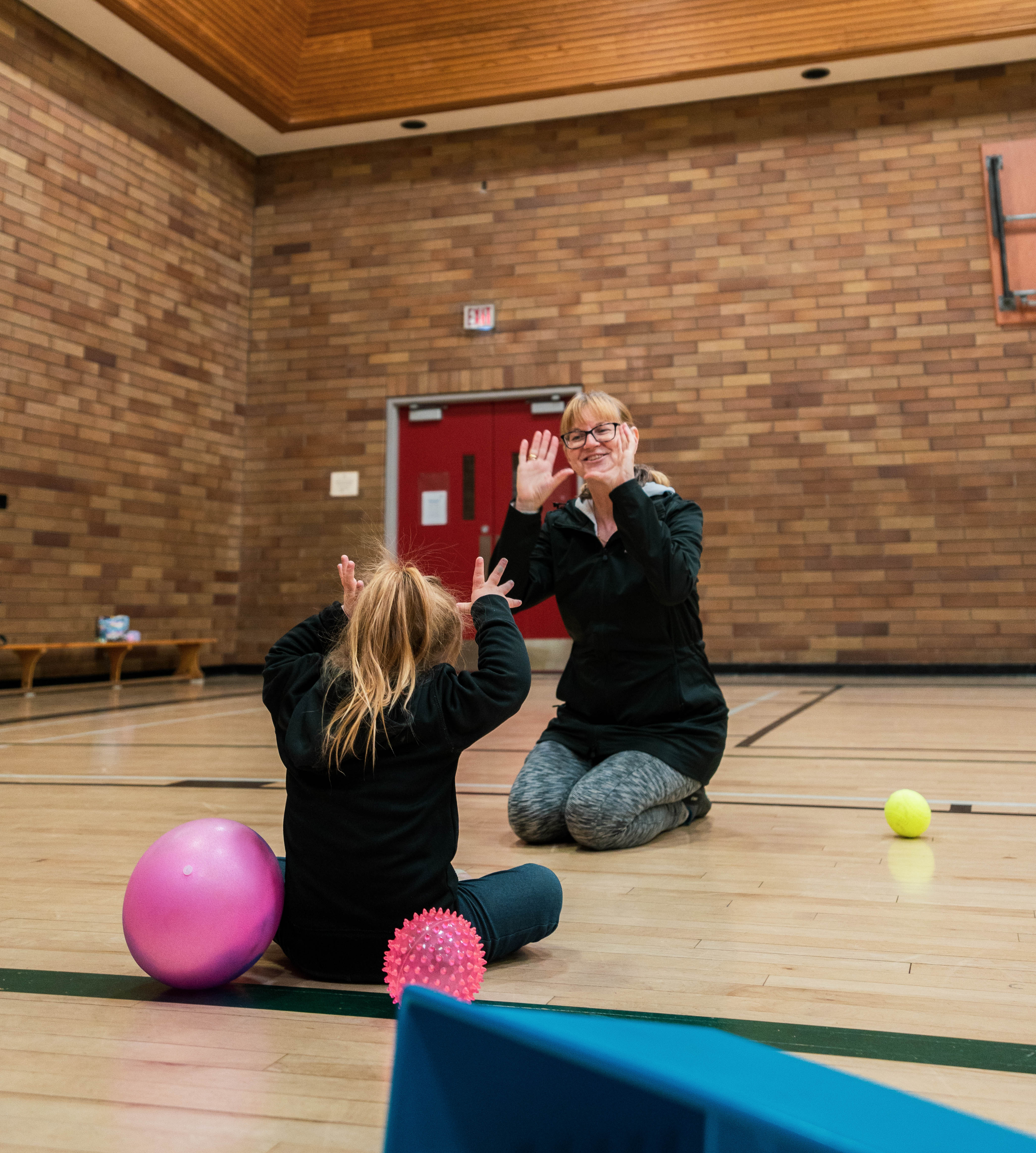 A young girl with her back to the camera is making gestures with her hands in play with her mom who sits opposite her. A young girl with her back to the camera is making gestures with her hands in play with her mom who sits opposite her.