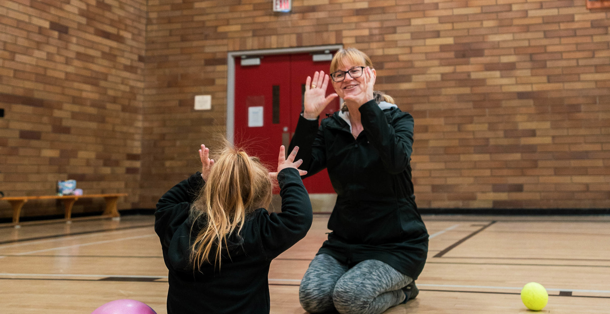 A young girl with her back to the camera is making gestures with her hands in play with her mom who sits opposite her.