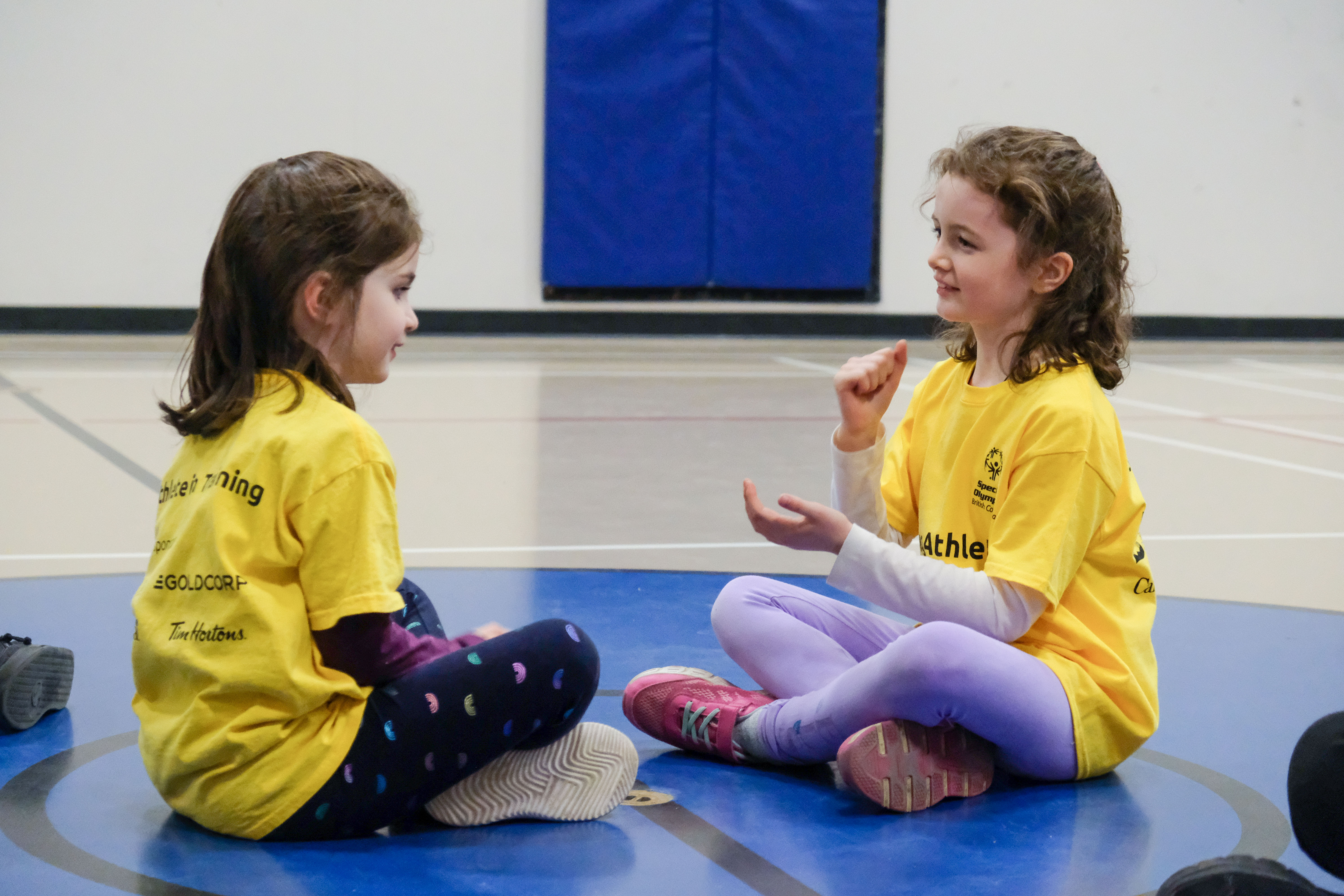 Two young girls with intellectual disabilities sit together wearing matching yellow shirts and playing a game with their hands. Two young girls with intellectual disabilities sit together wearing matching yellow shirts and playing a game with their hands.