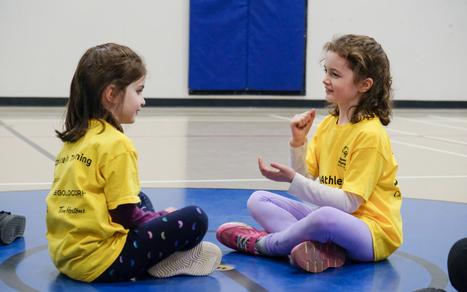 Two young girls with intellectual disabilities sit together wearing matching yellow shirts and playing a game with their hands.