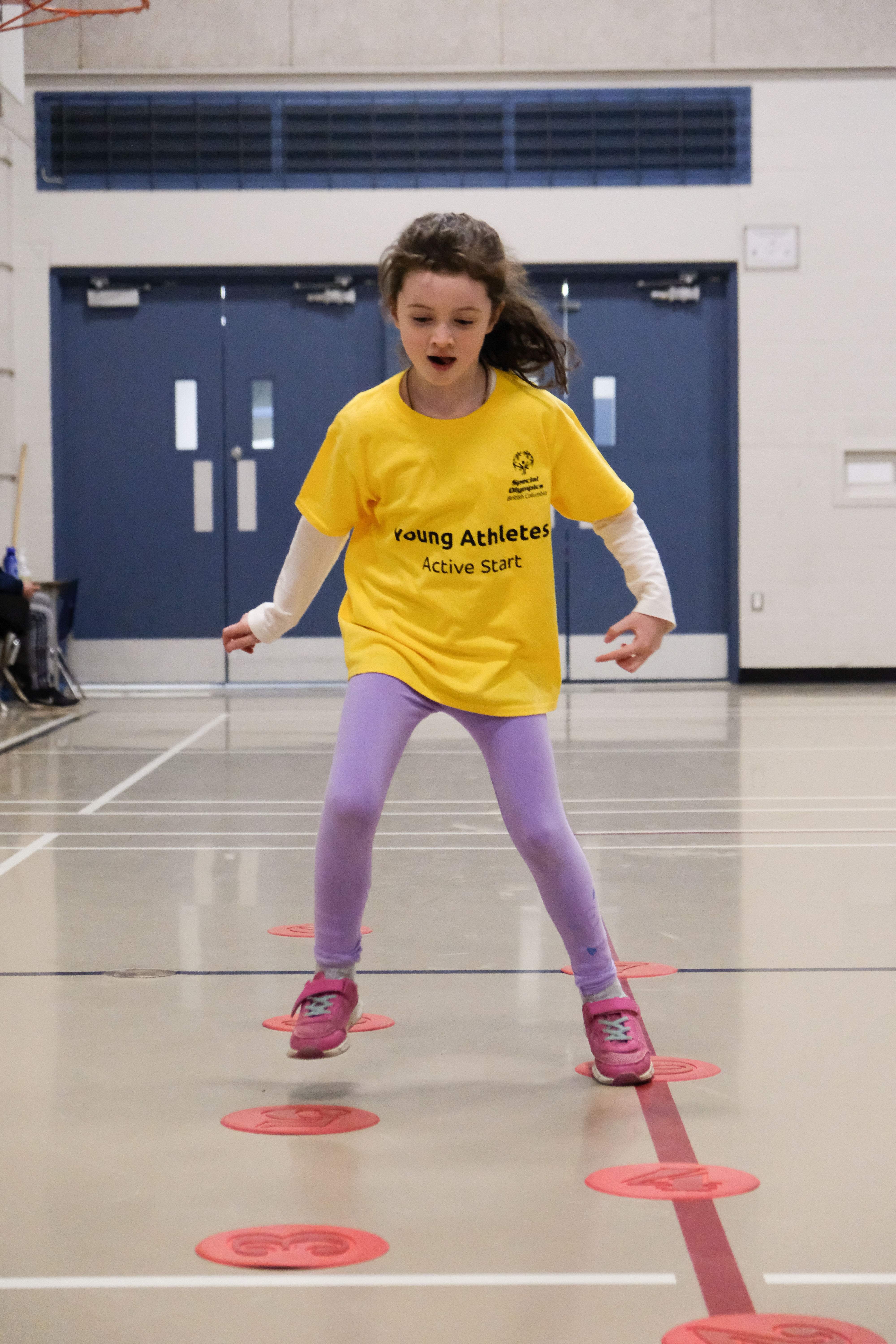 A young girl with intellectual disabilities jumps between markers on the floor looking very focused. A young girl with intellectual disabilities jumps between markers on the floor looking very focused.