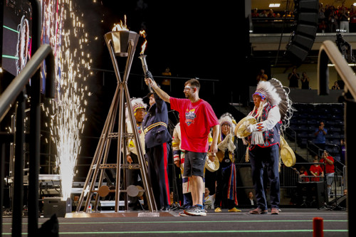 Law enforcement member and athlete holding up a flame to a cauldron, on stage with Indigenous performers.