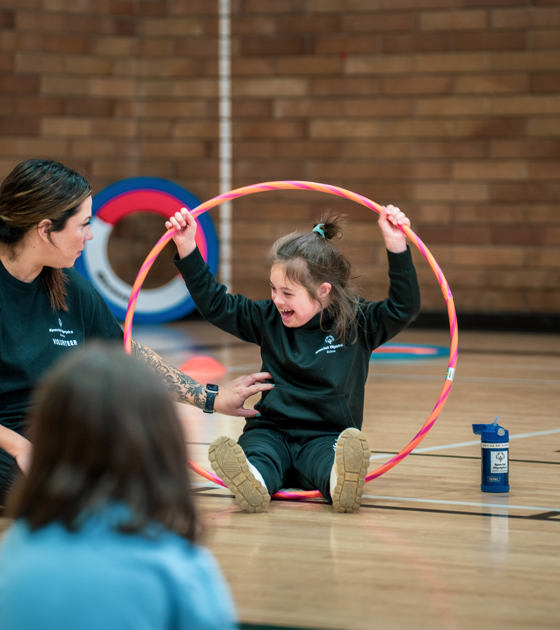 A young girl with intellectual disabilities sits on the floor of gym holding a large, pink hula-hoop over her head while interacting with her leader.