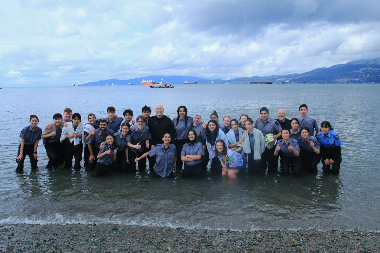 A large group of participants pose together in the shallow waters of English Bay looking proud. A large group of participants pose together in the shallow waters of English Bay looking proud.