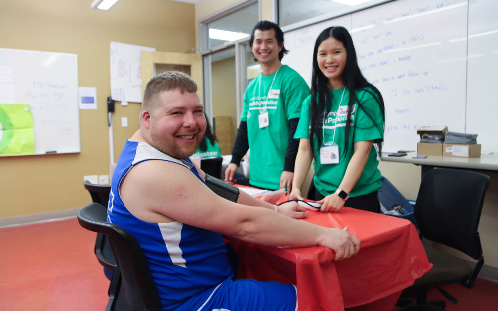 An athlete sits at a table getting his blood pressure taken by volunteers behind him.